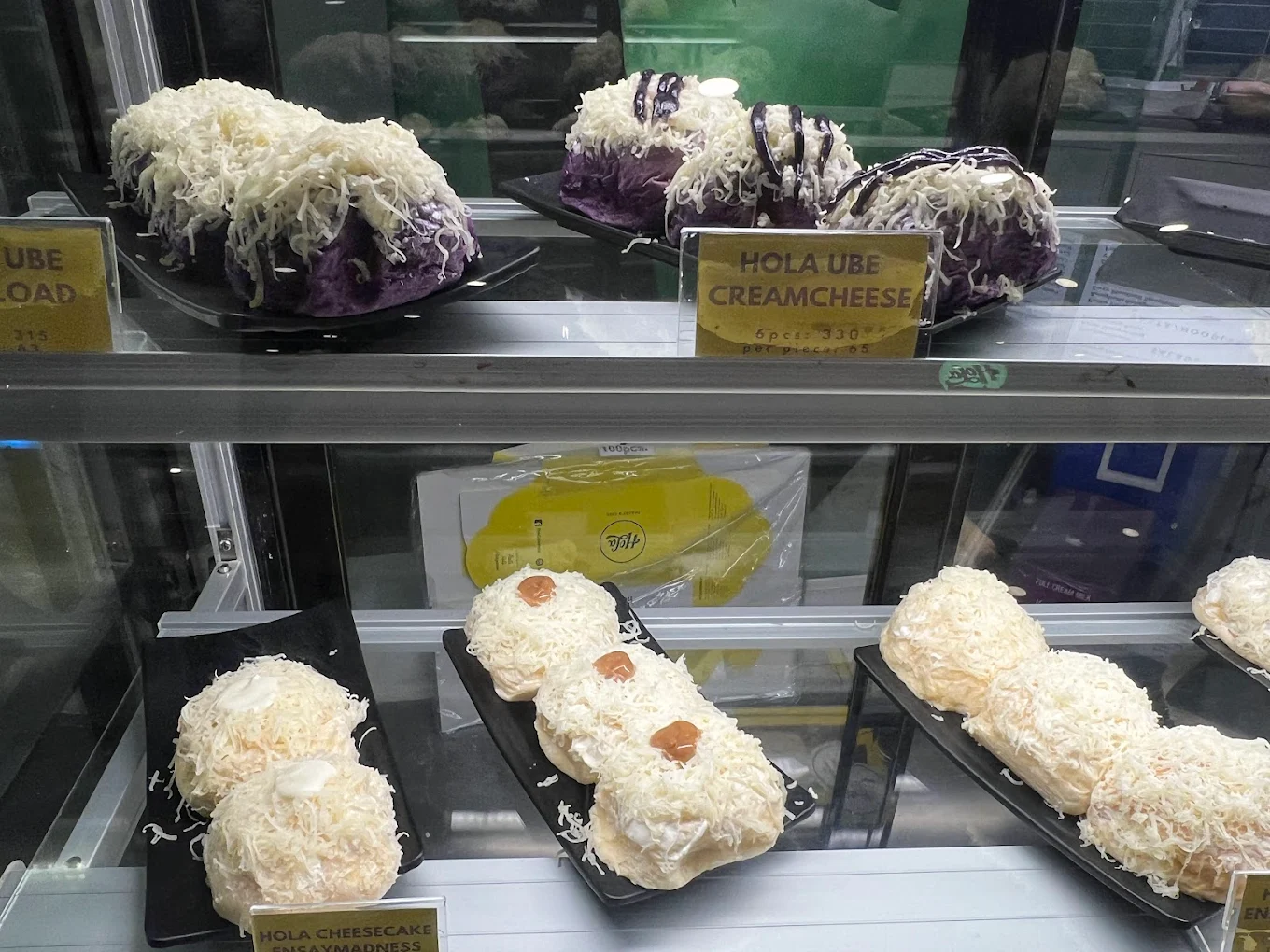 Hola Cream Cheese Ensaymadness in a bakery display case — pillowy ensaymada rolls smothered in grated cheese alongside Ube Cream Cheese ensaymada