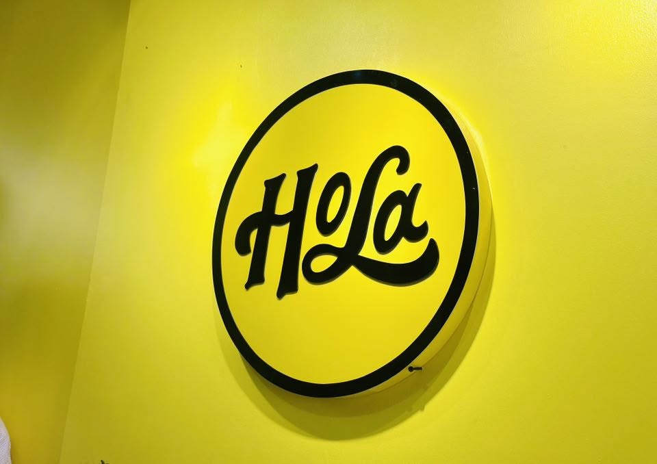 The Hola circle mark mounted on the signature sunrise-yellow wall inside a Cafe Hola branch